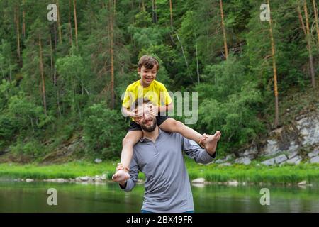 Happy father piggyback his son outside. Laughing boy closed his fathers eyes with his hands. Stock Photo