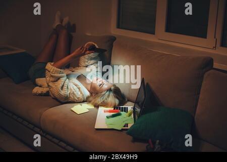 Tired caucasian student chatting on phone while sitting on sofa after doing homework late evening Stock Photo