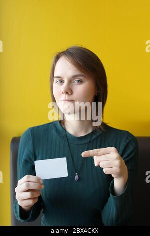 A young beautiful girl of European appearance in a blue jacket sits in a cafe on the couch against the background of a yellow wall. Pointing finger to blank white plastic card. Layout for design, copy space and place for text. Stock Photo