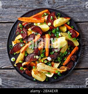 roasted sliced beetroot, potato wedges, parsnips, carrots sprinkled with chopped green onion on a black plate on a wooden table, horizontal view from Stock Photo