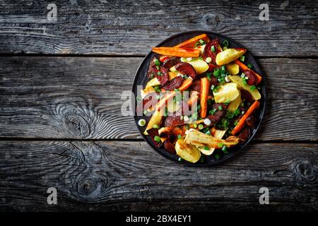 vegetables roasted in the oven, sliced beetroot, potato wedges, parsnip, carrots sprinkled with chopped green onion on a black plate on a wooden table Stock Photo
