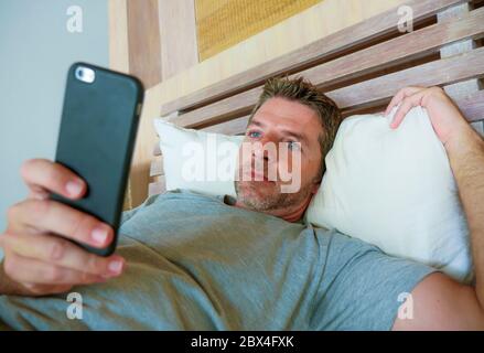 lifestyle indoors portrait of young happy and attractive man at home bedroom using internet social media app networking from bed thoughtful and relaxe Stock Photo