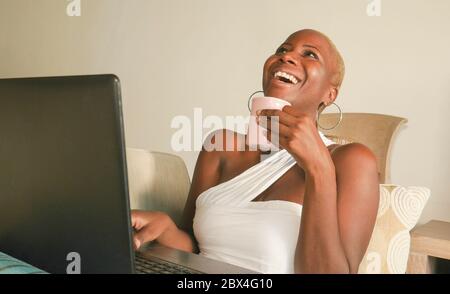 young beautiful and happy black afro American woman smiling excited having fun on internet using social media on laptop computer at home living room c Stock Photo