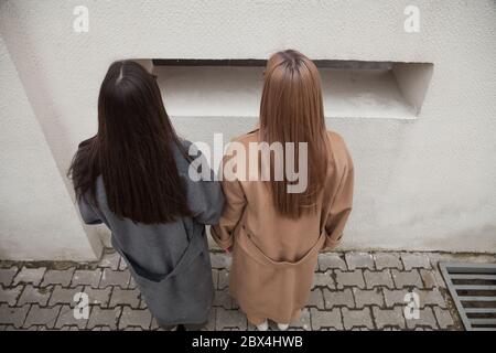 unrecognizable two models, backside view of two girlfriends, blonde and brunette in warm outwear camel and grey  coat over white wall background Stock Photo