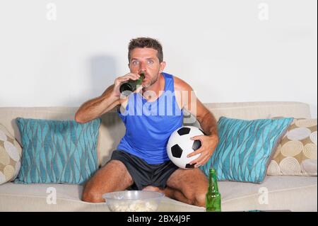 passionate football fan supporter man watching match on television drinking beer eating popcorn holding soccer ball sitting at home sofa couch nervous Stock Photo