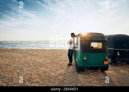 Tuk tuk driver waiting for passenger against sand beach and sea. Unawatuna, Sri Lanka. Stock Photo