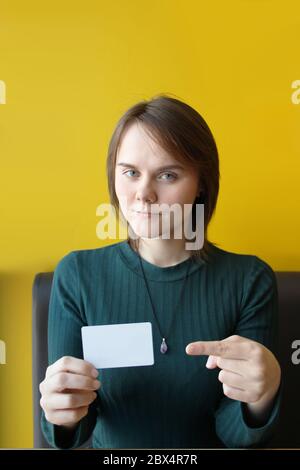 A young beautiful girl of European appearance in a blue jacket sits in a cafe on the couch against the background of a yellow wall. Pointing finger to blank white plastic card. Layout for design, copy space and place for text. Stock Photo