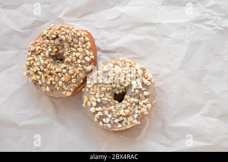 Brown glazed donuts sprinkled with nuts on crumpled beige paper background. Unhealthy high-calorie food. Empty place for text. Copy space Stock Photo