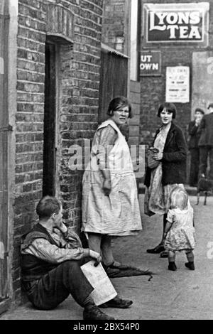 An English working class family in an alley in an English working class neighbourhood in London. The father is sitting with a newspaper leaning against the wall of a house, two women and a little girl in dresses are standing next to him. In the background there is a 'Lyons Tea' shop. Taken by Alfred Eisenstaedt during the European depression. Stock Photo