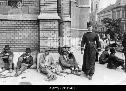 A field chaplain of the Imperial and Royal Army, in his uniform, crosses a square where many peasants sit bored on the ground. The Imperial and Royal Army is the joint army of the Austro-Hungarian Dual Monarchy. Stock Photo
