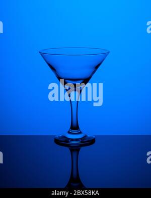 Glass goblet without wine on a thin leg stands on a mirror surface. Back light. Dark blue background. Vertical orientation. Stock Photo