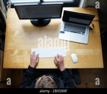 Top shot of real male web programmer coding javascript, php, symfony, react, working with keyboard and mouse with laptop and external monitor from hom Stock Photo