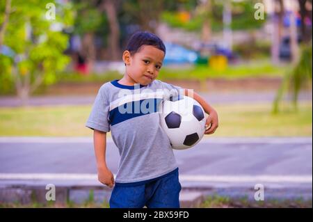 lifestyle outdoors portrait of 5 or 6 years old young Asian Indonesian kid posing happy with soccer ball playing football in child sport practice educ Stock Photo