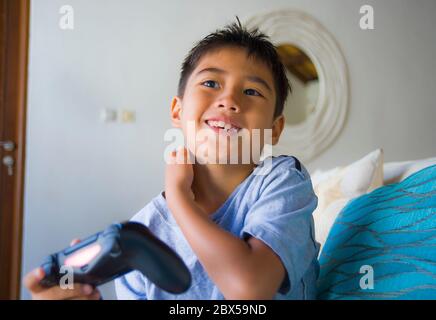 lifestyle portrait of Latin young kid 8 years old excited and happy playing video game online holding remote controller enjoying having fun on couch i Stock Photo