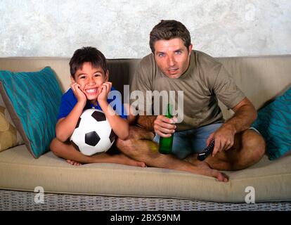 lifestyle portrait of father and little boy watching football game together on television at home living room couch crazy excited holding ball in fath Stock Photo