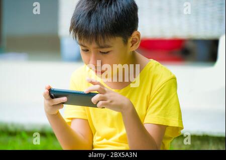 isolated lifestyle portrait of 7 or 8 years old Asian child focused and concentrated playing with mobile phone outdoors at home garden in kid sufferin Stock Photo