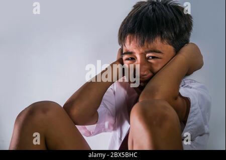 lifestyle dramatic bullying victim portrait of young sad and scared hispanic kid 7 - 10 years old in school uniform sitting alone crying depressed and Stock Photo