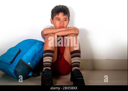 lifestyle dramatic bullying victim portrait of young sad and scared hispanic kid 7 - 10 years old in school uniform sitting alone crying depressed and Stock Photo