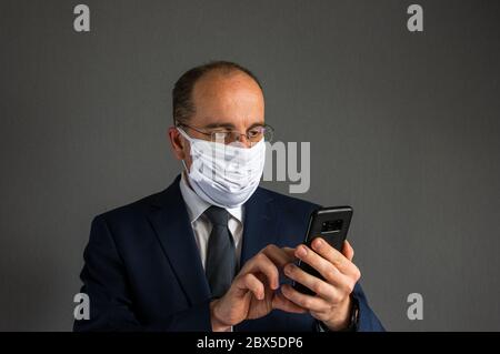 portrait of a young business man wearing a suit and tie and protective face mask checks information on his smartphone in front of gray background. Lot Stock Photo