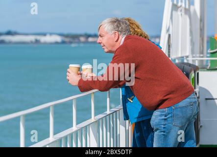 Middle aged couple holding cups and drinking takeaway coffee while looking over the railings on a ferry on the sea. Stock Photo
