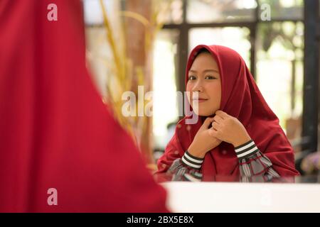 mirror reflection of young happy and beautiful Muslim woman in traditional hijab adjusting her red head scarf  cheerful dolling up in beauty and fashi Stock Photo