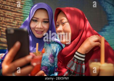 Muslim woman and her teenager daughter . Happy Asian Indonesian girl taking selfie with her beautiful mum both in traditional Islamic hijab head scarf Stock Photo