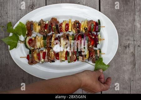 Skewer before grilling composed of bacon onions tomatoes meat pork zucchini peppers on a plate with leaves and hand on a wooden table Stock Photo