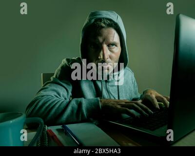dangerous looking young hacker man in hoodie typing on laptop computer hacking and decoding system data or having illegal access breaking password on Stock Photo