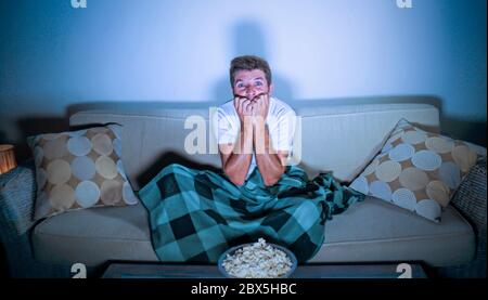 lifestyle portrait of attractive scared and nervous man watching suspense horror movie on television feeling stressed covering with blanket in panic s Stock Photo