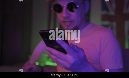Hookah smoker using mobile phone. Handsome Caucasian Man in sunglasses sits in neon backlit. Selective focus on male hand holding phone in foreground Stock Photo