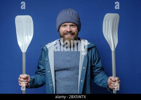 Man in a hat and a raincoat holds oars in hands, he got ready for the long-awaited travel and is very happy. Blue background. Stock Photo