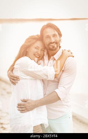 Happy couple hugs on the beach. Smiling man and woman in white clothes looking at camera while standing embracing on sea coast against backdrop of Stock Photo