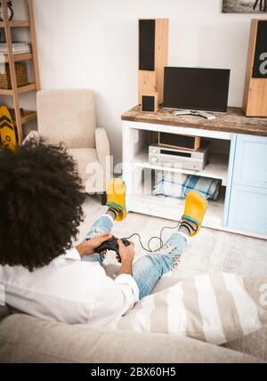Young man playing computer games sitting at home on couch in front of TV. Rear view of unrecognizable gamer removing buttons on Play Station joystick Stock Photo