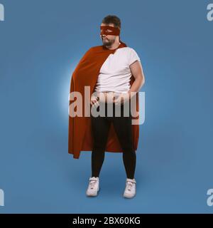Body positive Super hero man added weight over winter. Sad man in superhero costume checks fat on his tummy. Isolated on blue background. Template Stock Photo