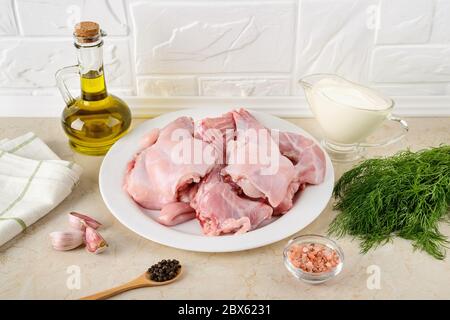 Pieces of raw rabbit meat, sour cream, garlic, pepper, salt, olive oil and bunch of dill on kitchen table. Ingredients for cooking rabbit stew. Stock Photo