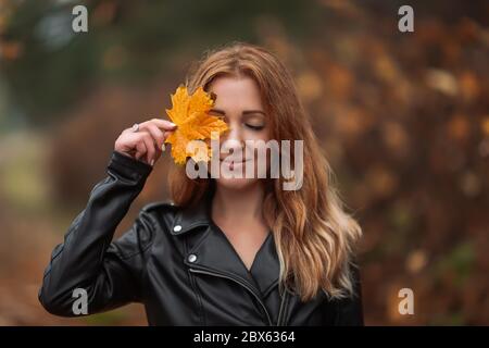 Portrait of smiling redhead woman with yellow leaf with long curly hair on blurred autumn background. Girl on fabulous background of park with orange Stock Photo