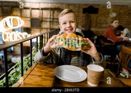 Happy cute boy sitting at the table in city cafe, smiling to camera and holding fresh croissant with salad and vegetables. Tasty lunch in cafe Stock Photo