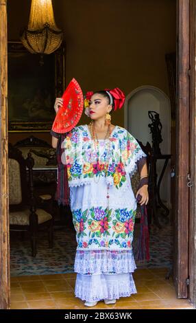 Merida,Yucatan/Mexico-February 29,2020: woman in traditional embroidered huipil tunic and dress stands with her red fan in hacienda's doorway Stock Photo