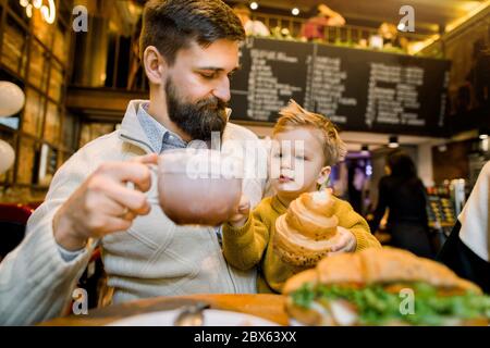 Happy handsome bearded Caucasian man, father, sitting at the table with his little son and holsing cup of latte or cocoa drink. Little cute boy Stock Photo