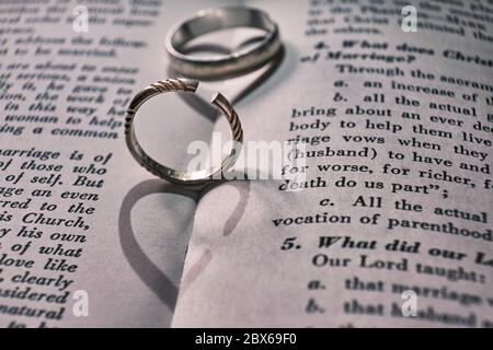 Two wedding bands, one has been cut off on a book with text about marriage. The shadows from the rings make the shape of a heart, and a broken heart. Stock Photo