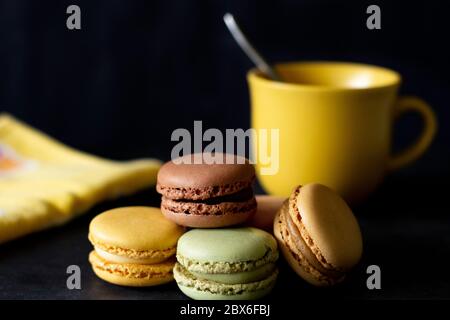 Group of colored macarons with unfocused yellow cup and napkin on black background. Stock Photo