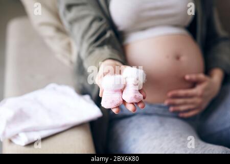 Hand of young pregnant woman holding two tiny pink woolen socks in front of camera while sitting on couch and touching her belly Stock Photo