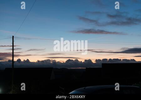 Telegraph pole and lines, low buildings and dark cumulus clouds silhouetted against pale blue and orange evening sky. Stock Photo