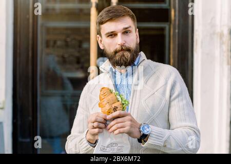 Young handsome bearded man posing in front of cafe door in the street holding his croissant with lettuce and fresh vegetables. Cafe concept. Healthy Stock Photo