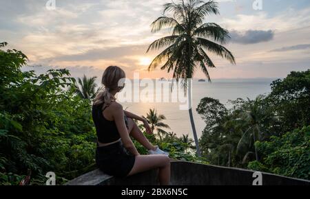 Silhouette of woman sitting alone on the hill with a coconut palm trees and enjoys sea on a horizon in a sunset. Vacation and travel concept on a Stock Photo