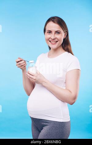 Pretty pregnant woman in activewear holding small plastic jar with bio yoghurt while eating it in front of camera in isolation Stock Photo