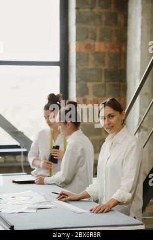Young confident designer of clothes looking at you while standing by table on background of two colleagues discussing new sketches Stock Photo