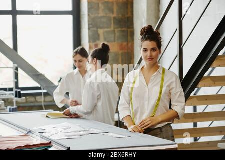 Young serious female tailor with measuring tape on her neck standing by table in workshop with her colleagues discussing ideas near by Stock Photo