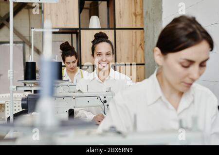 Happy young seamstress with toothy smile looking at you among her colleagues while sewing new clothes by machine in workshop Stock Photo