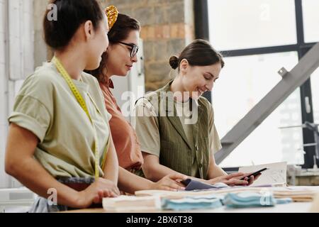 Happy young fashion designer with smartphone showing her colleagues some new items for their seasonal collection and discussing them Stock Photo